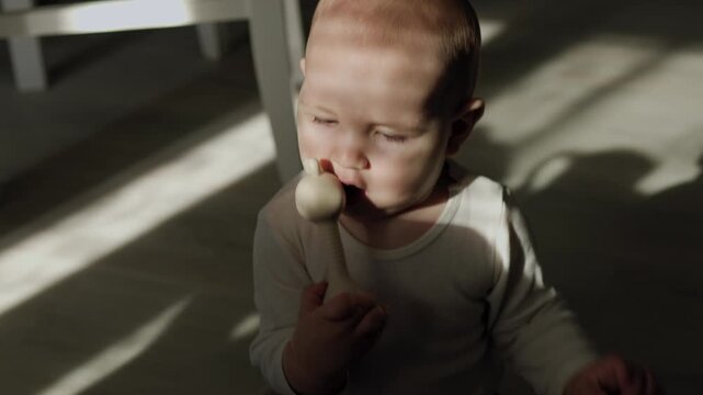 Infant in the teething phase crawling on an indoor floor and biting a silicone teether. Strong sunlight casts distinct shadows across the baby's face and body during daytime.