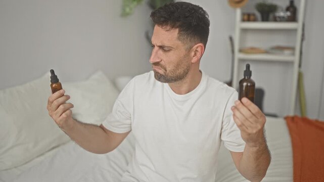 Man young hispanic in white t shirt holds two amber dropper bottles in each hand comparing labels while seated on a bed in a bedroom; doubt choice grooming.