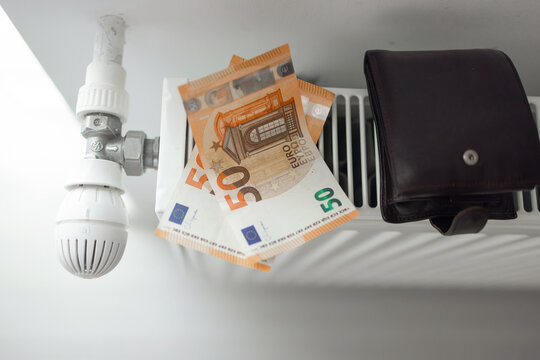 Hands adjusting a white radiator thermostat and feeling the heater. The set includes 50 Euro banknotes and a black wallet placed on the heating equipment.