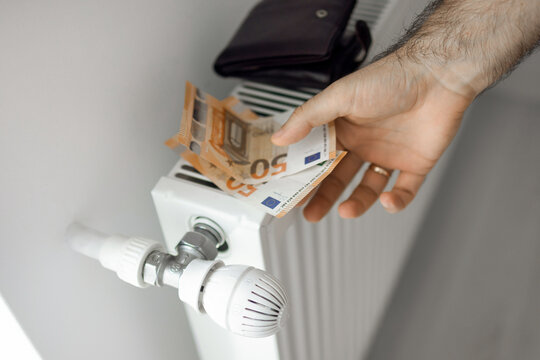 Hands adjusting a white radiator thermostat and feeling the heater. The set includes 50 Euro banknotes and a black wallet placed on the heating equipment.
