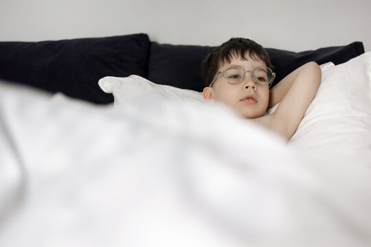 Clips of a young boy wearing glasses playing and relaxing in a bed with a white blanket. He wraps himself in the duvet, smiles, and rests with his feet visible under the covers.