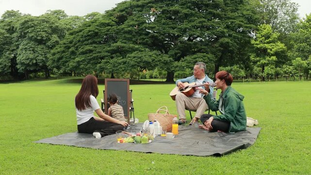 Asian kids painting in park. Asian family enjoy outdoor picnic with food and drinks in park sunny day, outdoor activitie with daughter father, mother grandfather. GrandFather playing acoustic guitar