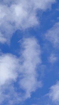 A small rocket flies high through a bright blue sky surrounded by soft white clouds. The distant flame, altitude, and open atmosphere create a dramatic aerospace launch scene.