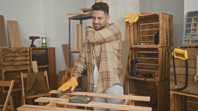 Man carpenter wearing yellow gloves sanding a plank with a handheld sander, rubbing shoulder and wincing amid wooden crates and power tools in studio; fatigue.