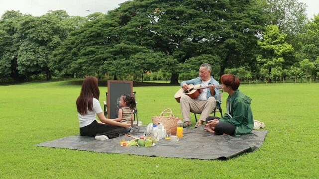 Asian kids painting in park. Asian family enjoy outdoor picnic with food and drinks in park sunny day, outdoor activitie with daughter father, mother grandfather. GrandFather playing acoustic guitar