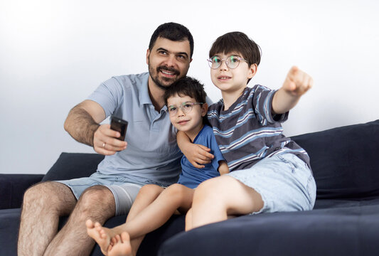 Father and his two sons sitting on a couch and watching television in a living room. The man holds a remote control while relaxing with the boys indoors