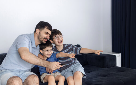 Father and his two sons sitting on a couch and watching television in a living room. The man holds a remote control while relaxing with the boys indoors