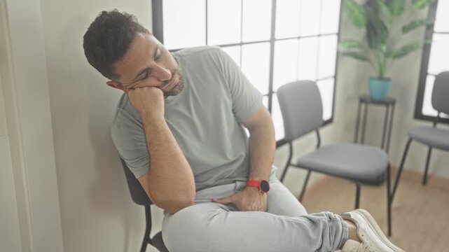 Man resting head on hand, sitting in chair by bright window, wearing gray tshirt, jeans and white sneakers in a clinic building waiting area; boredom fatigue.