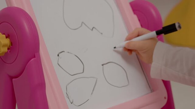 A young child happily uses a pink drawing board to create simple shapes. The colorful setting encourages creativity as they focus on their artwork.