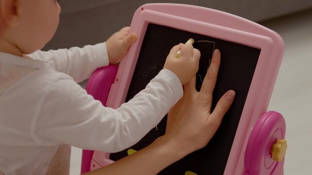 A young boy skillfully outlines his mother's hand using yellow chalk on a pink drawing board in a cozy indoor setting while she guides him gently..
