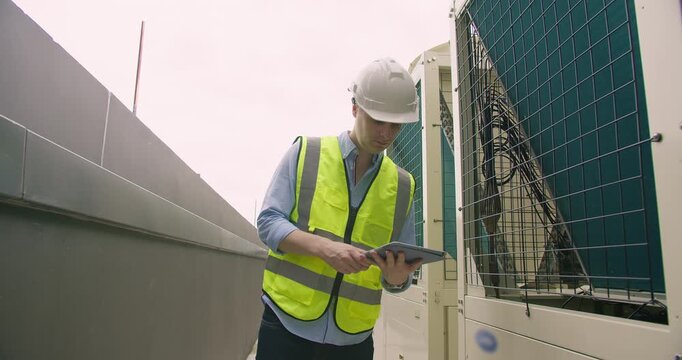 A professional maintenance technician wearing a safety vest inspects an industrial HVAC unit on a rooftop, using a tablet for system diagnostics and quality control.