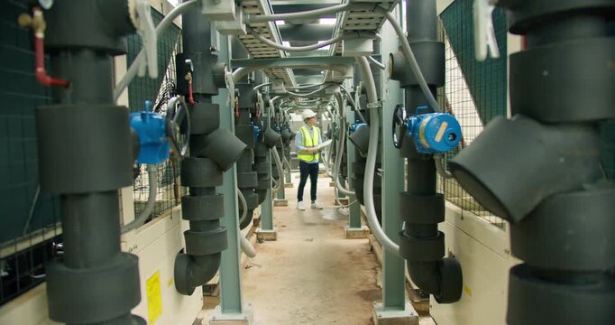 An engineer in a hard hat inspects an industrial HVAC system's pipes and valves, performing a routine maintenance check on a large-scale commercial building rooftop.