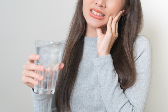 Suffering from toothache, hurt asian young woman touch cheek, face expression ache or feel pain, sensitive molar teeth, hand holding glass of water with ice, inflammation when drink cold, healthcare.