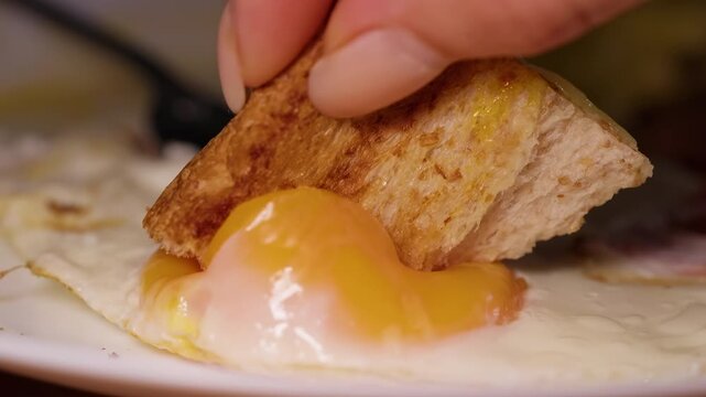 Hand Dips Toasted Bread Into a Runny Fried Egg Yolk on a White Plate, Close Up Food Preparation and Breakfast Meal