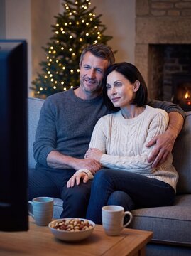 A middle-aged couple sits together on a couch watching television during the holiday season, surrounded by warm lights from a decorated Christmas tree and a fireplace in a cozy living room.