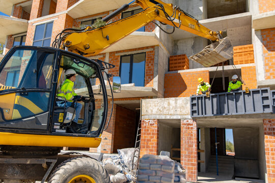 Woman operating wheeled excavator while team lifts metal formwork