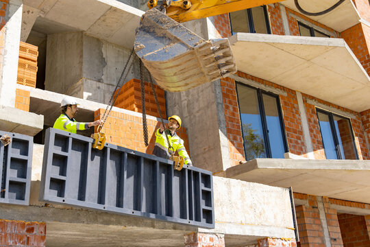 Builders guiding suspended metal panel lifted by excavator bucket