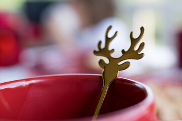 Macro shot focusing on a brass reindeer head-shaped drink stirrer resting in the rim of a red ceramic mug during a festive holiday breakfast. © Phoscar