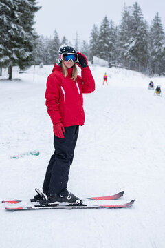 A young girl adjusts her goggles while standing on skis at a snowy winter resort. The scene represents ski preparation, youth activity, and winter sports learning.