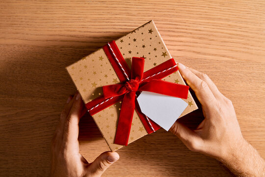 Hands Holding Beautifully Wrapped Christmas Gift Box with Red Velvet Ribbon on Wooden Table