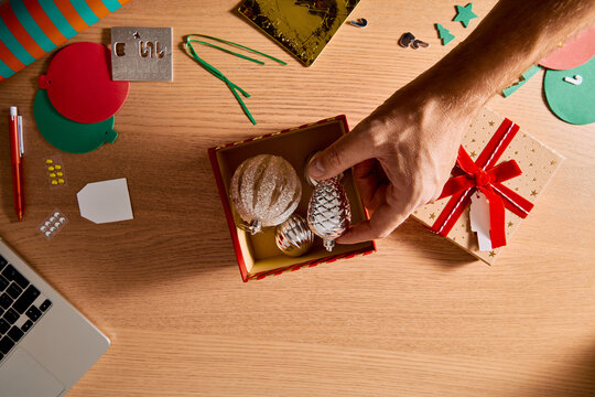 Person Packing Christmas Ornaments into Gift Box on Wooden Desk with Holiday Craft Supplies