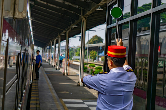 A station master complete with his uniform and red hat is holding up the green sign while checking his watch, indicating that the train is allowed to depart