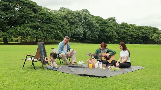 Asian kids painting in park. Asian family enjoy outdoor picnic with food and drinks in park sunny day, outdoor activitie with daughter father, mother grandfather. GrandFather playing acoustic guitar