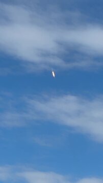 A rocket climbs through a bright blue sky with soft white clouds after liftoff. The glowing flame, vertical ascent, and open atmosphere create a dramatic aerospace launch scene.