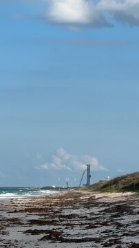 A rocket booster descends through the blue sky near a coastal launch pad. The beach, dunes, smoke, and landing flame show a dramatic spaceflight recovery scene.