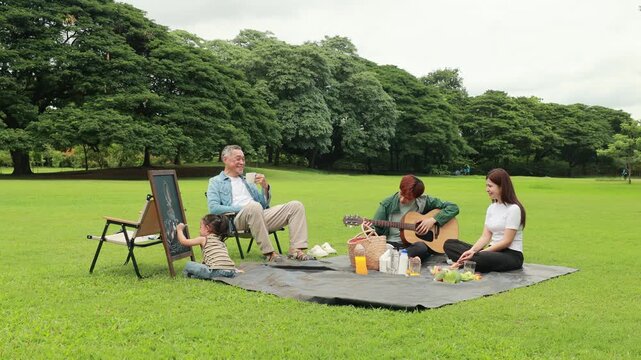 Asian kids painting in park. Asian family enjoy outdoor picnic with food and drinks in park sunny day, outdoor activitie with daughter father, mother grandfather. GrandFather playing acoustic guitar
