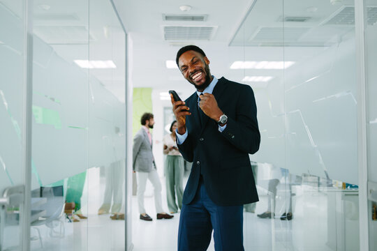 Happy african american businessman celebrating success with a mobile phone in a modern office hallway, receiving good news and achieving a business goal