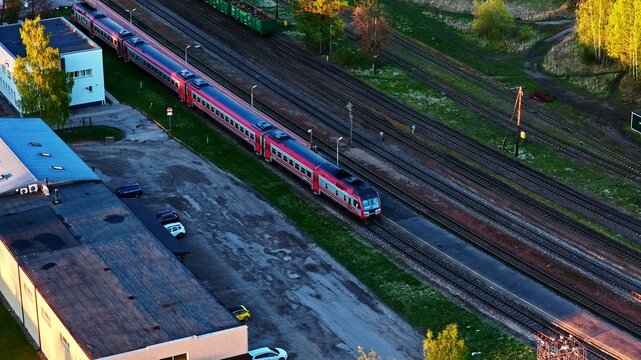 Aerial view of a passenger train traveling along railway tracks next to industrial buildings and green trees in Latvia.