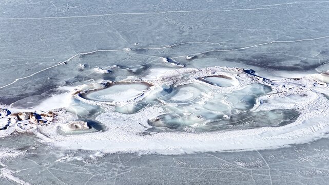 Aerial view of frozen lake surface with intricate ice patterns, cracks, and patches of open water in Latvia.