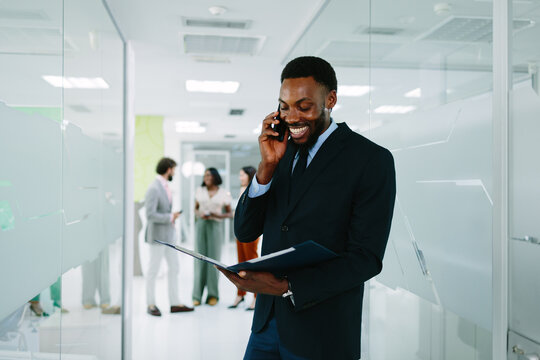 Happy black businessman communicating on smartphone, holding documents, walking through modern office corridor, networking with diverse team in background