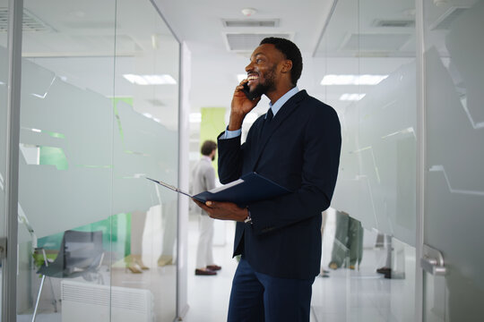 African american businessman communicating on smartphone, walking through modern corporate office hallway, holding a folder, smiling, and feeling confident about success