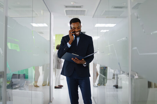 Black businessman walking a glass-walled office corridor, smiling and talking on his phone while carrying a document folder, conveying confidence, productivity and positivity