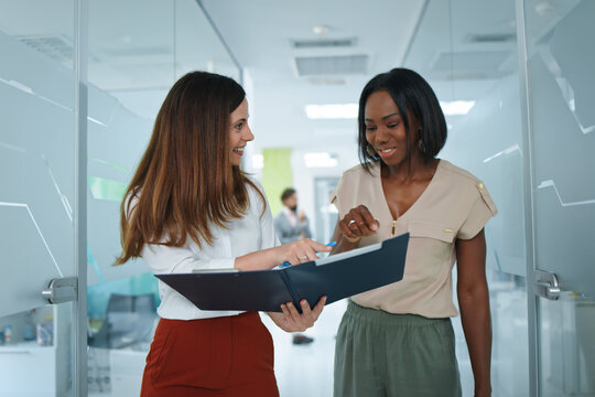 Diverse businesswomen collaborating on a project, discussing documents in a modern office hallway, sharing ideas and expertise for successful teamwork and professional communication