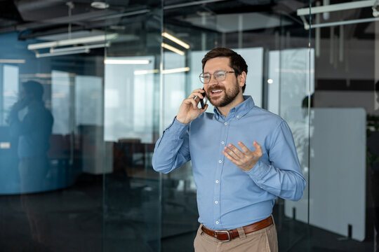 A smiling young businessman in a shirt and glasses stands in the office, looking out the window and talking on the phone while gesturing with his hand