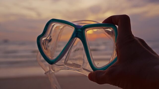 Hand Holds Teal Diving Mask Before Ocean Sunset on Sandy Beach, Evoking Summer Travel and Water Recreation