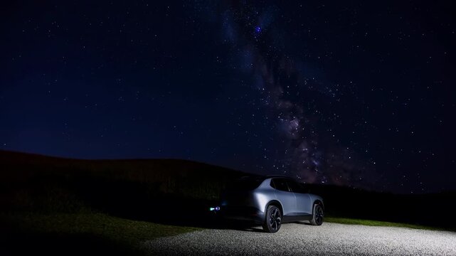 Silver SUV remains parked on gravel roadside beneath star-filled night sky and bright Milky Way, illuminated against dark rolling hills with wide cinematic landscape composition