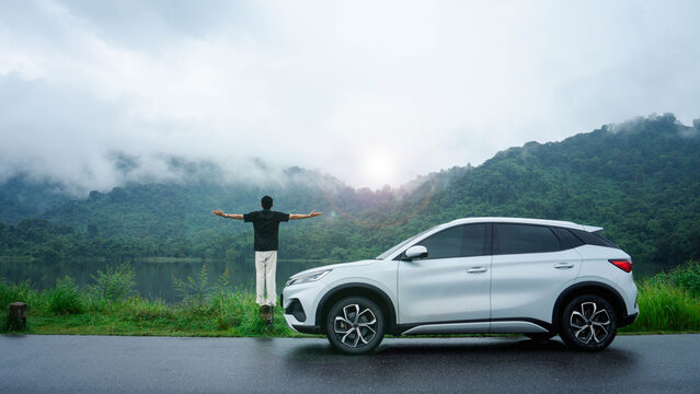 Man spreads his arms beside a car overlooking a misty mountain lake. The scenic road trip represents travel freedom, outdoor adventure, relaxation, nature tourism, and a carefree vacation lifestyle.