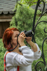 woman with red hair holds black digital camera near eye, profile, portrait, close-up, mirrorless, middle age, lens mounted, greenery in background, green plants, beautiful, romance, white blouse, take © Studio  GULL