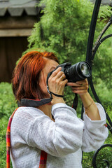 woman with red hair holds black digital camera near eye, profile, portrait, close-up, mirrorless, middle age, lens mounted, greenery in background, green plants, beautiful, romance, white blouse, take © Studio  GULL