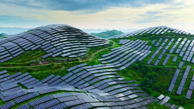 Vast arrays of photovoltaic solar panels cover rolling green mountain slopes under a bright blue sky filled with clouds during daytime.