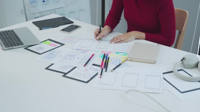 A focused female designer in a red sweater sketches mobile app wireframes with colored pens at a bright office desk surrounded by tablets a laptop and sticky notes while planning user interface layout