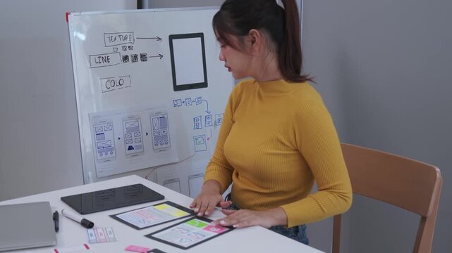A young woman in a yellow sweater arranges printed mobile app wireframes on a white table while glancing at a whiteboard filled with design sketches and workflow diagrams in a bright modern studio