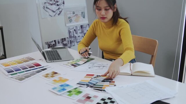 A young female interior designer sits at a white desk comparing color palettes and fabric swatches while referencing sketches and a laptop in a bright creative studio filled with architectural drawing