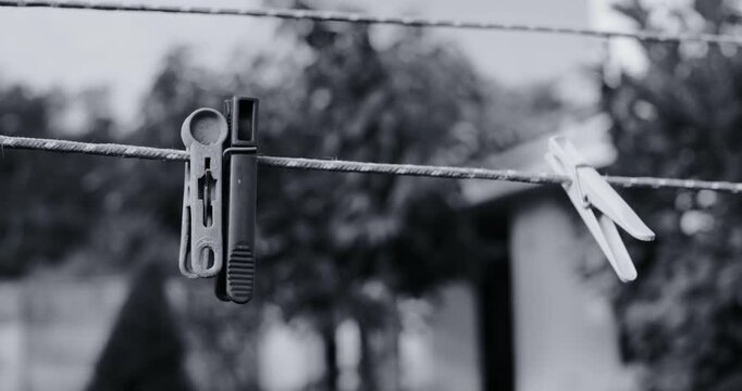Black and white photo of three clothespins hanging on a wire clothesline outdoors. Soft blurred background with garden plants and a fence. Vintage film grain style. Everyday rustic scene.