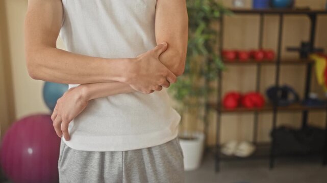 Young hispanic man holding left elbow with opposite hand in a fitness studio during yoga training, showing elbow pain; pain recovery.
