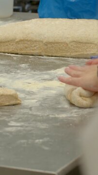 right hand of baker presses and guides small wholegrain dough portion on floured stainless steel worktop while larger rolled dough log rests in background